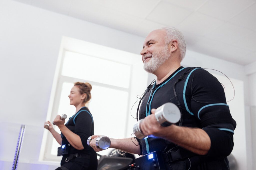 Man and woman wearing EMS suit for fat loss performing bicep curls with dumbbells during a workout session.
