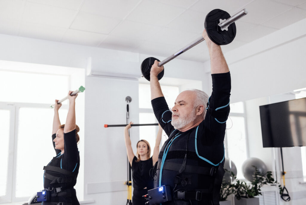 Group of older adults wearing EMS suit for fat loss and lifting barbells overhead during a guided fitness class