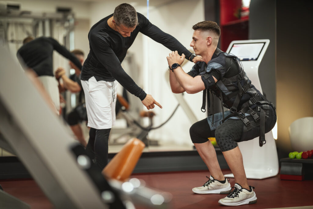 Man performing squats to demonstrate EMS training benefits while a trainer assists with proper form in a gym setting