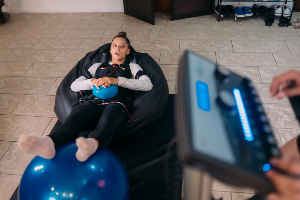 Woman reclining in an EMS training suit with an exercise ball, resting once she learned how long is EMS training actually takes.